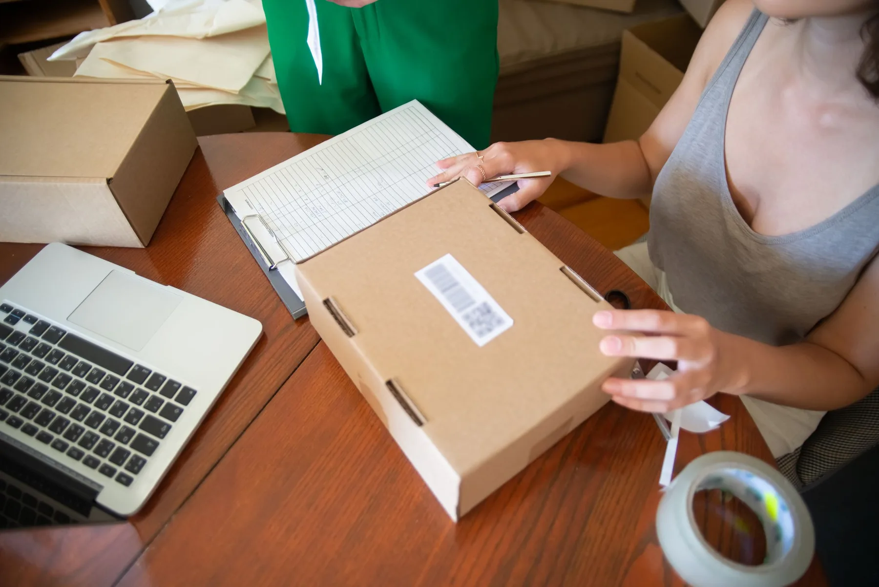 An ecommerce operator prepares a cardboard order beside a laptop and packing list.